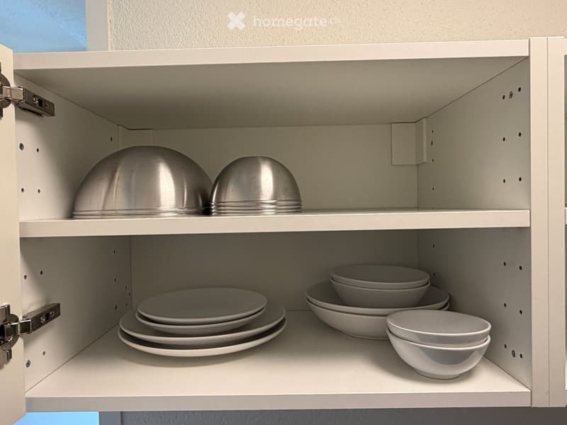 white cupboard with three shelves and two hanging rails, silver and white dishes, including plates, bowls, and pots
