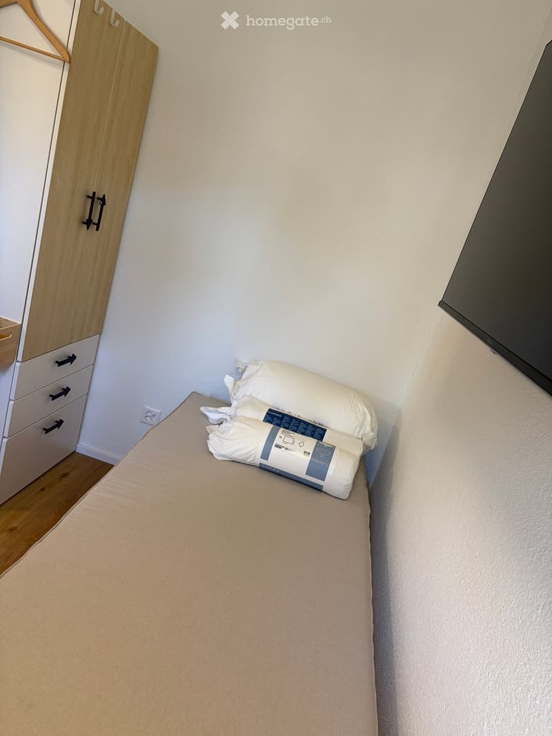 Small bed with two rolled up blankets on it, a wardrobe, a bedspread, and an electrical outlet