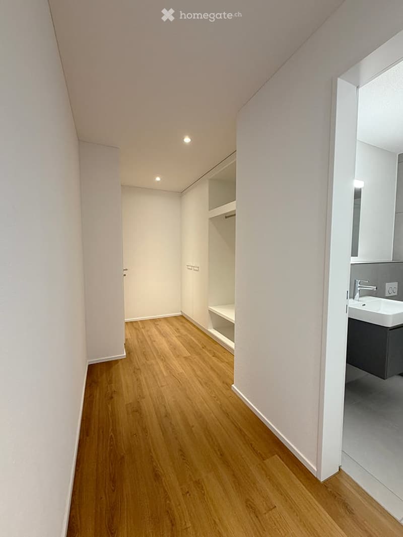 A narrow hallway with light wooden flooring, white walls, and recessed ceiling lighting. A built-in shelf unit is visible on one side, and an open doorway leads to a small bathroom with a sink and mirror.