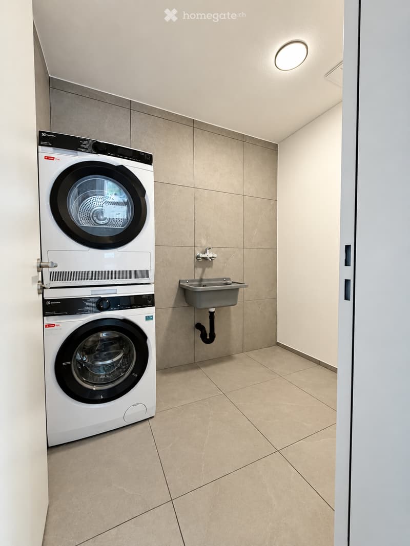 Modern laundry room with a stacked washer and dryer, a small sink, beige tiled walls and floor, and a round ceiling light.