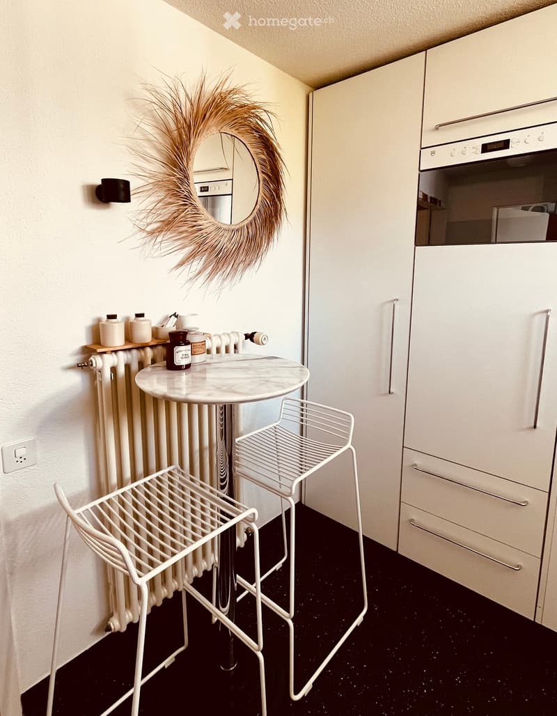A small, modern kitchen area with white cabinets, a marble table, two white metal chairs, a radiator, and a decorative mirror with a woven wreath.
