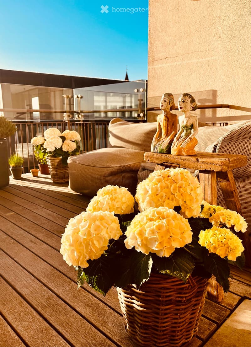 A wooden deck terrace with a sofa, small table with two decorative statues, and several flower arrangements in wicker baskets.