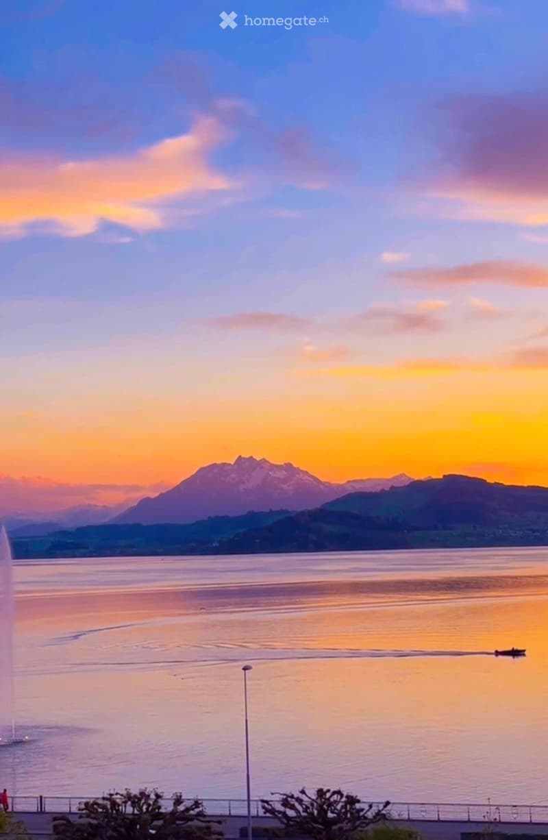 A scenic view of a lake at sunset with a fountain, mountains in the background, and a few people walking along the waterfront.