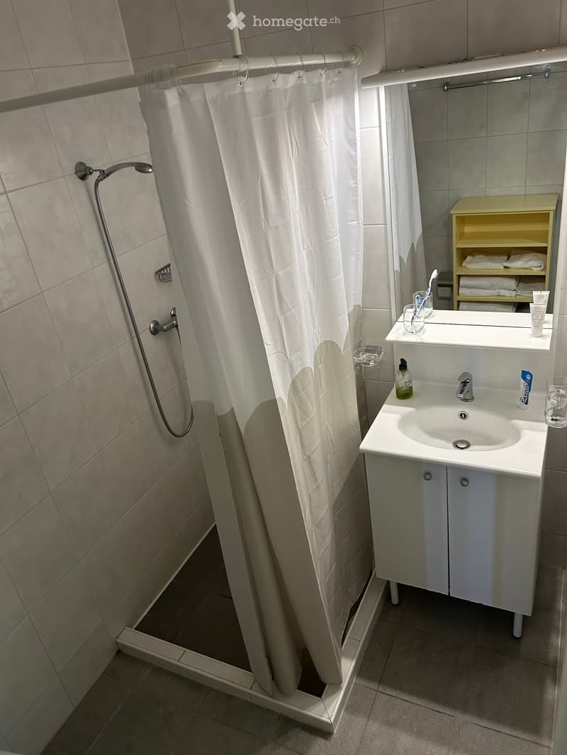 Modern bathroom with a white tiled shower area, a white sink with a mirror, and a small cabinet with towels.