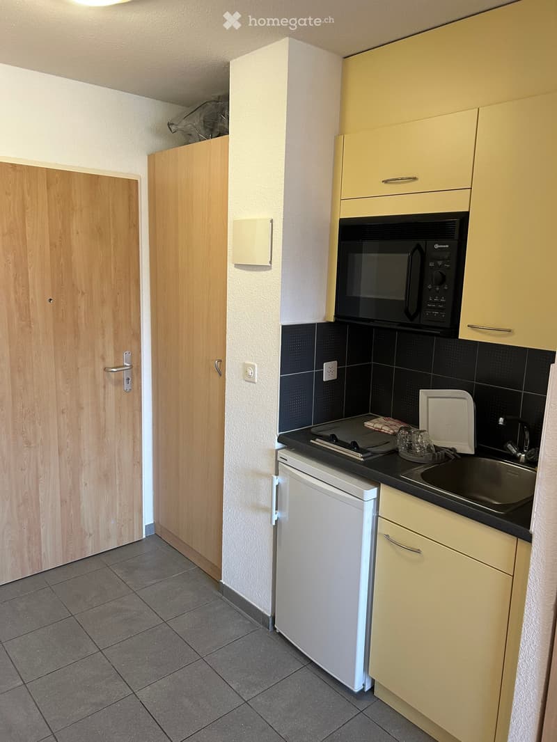 Compact kitchen with light-colored cabinets, a microwave, a small fridge, and a sink. The countertop is black, and the floor is tiled.