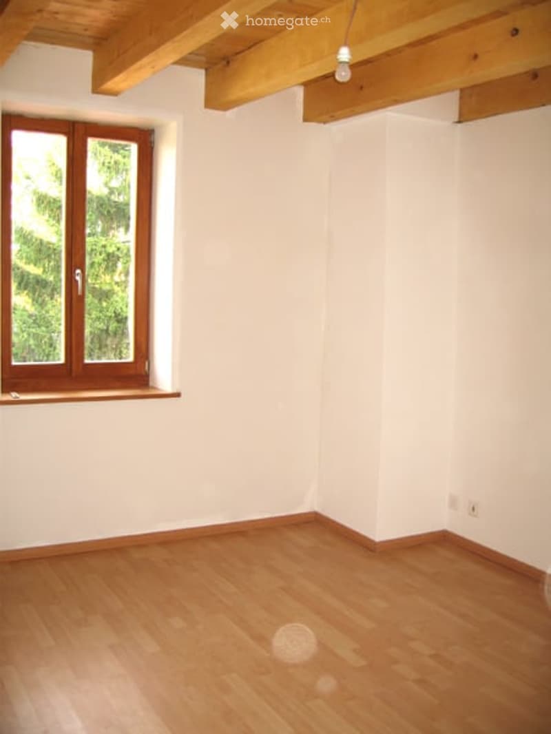 Empty room with wooden flooring, white walls, and exposed wooden ceiling beams. A single window with wooden frame and glass panes is visible on one wall.
