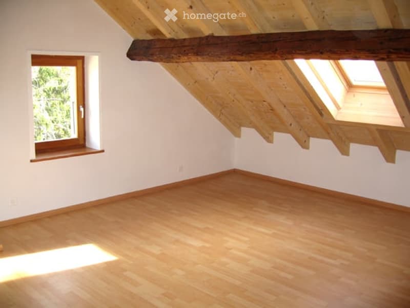 Empty attic room with wooden floors, white walls, exposed wooden beams, skylights, and a window with a wooden frame.