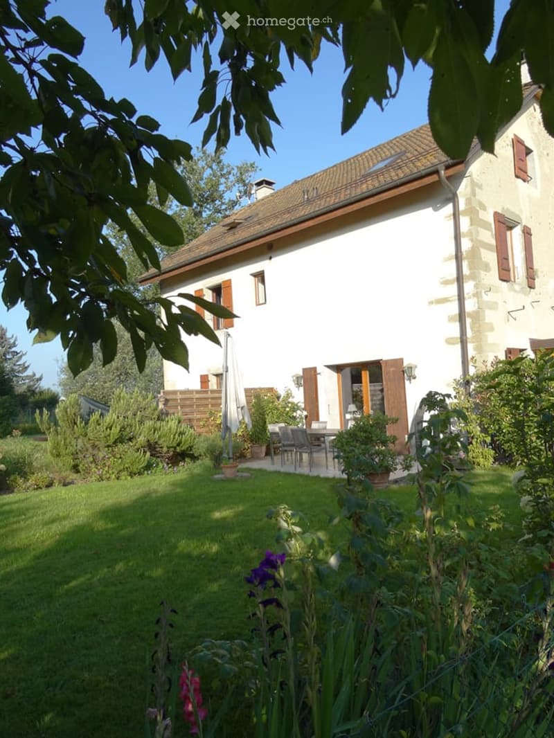 A well-maintained garden area in front of a two-story house with a tiled roof and wooden shutters. The garden features a neatly trimmed lawn, various potted plants, and a small outdoor seating area with a table and chairs.