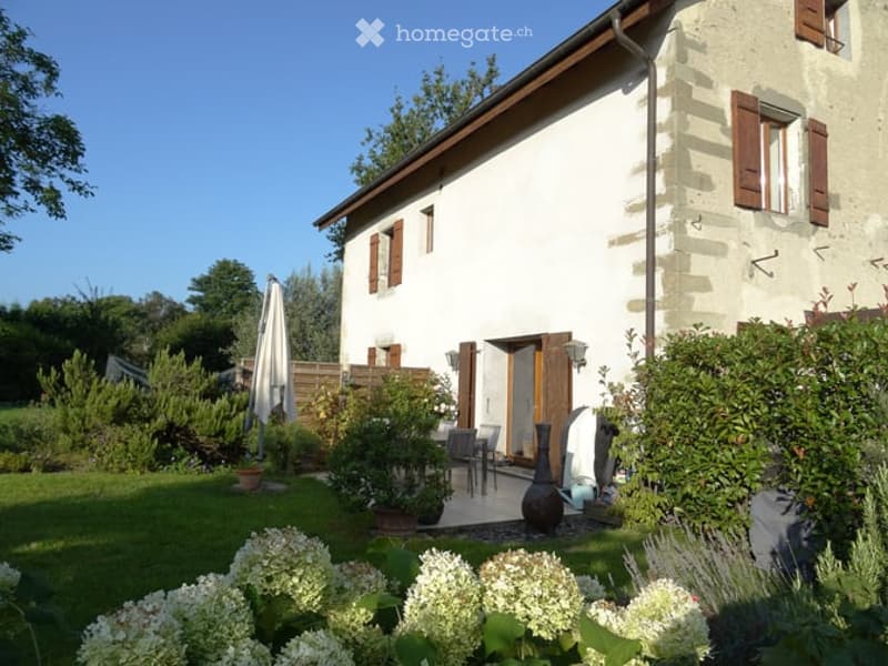 A charming countryside house with a well-maintained garden, featuring a patio area with outdoor furniture, potted plants, and a closed umbrella.