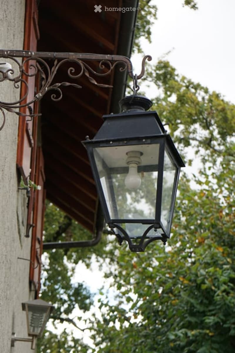 An exterior view of a building featuring a classic black lantern-style light fixture mounted on a decorative wrought iron bracket attached to the wall. The background includes a wooden roof overhang and lush green foliage.