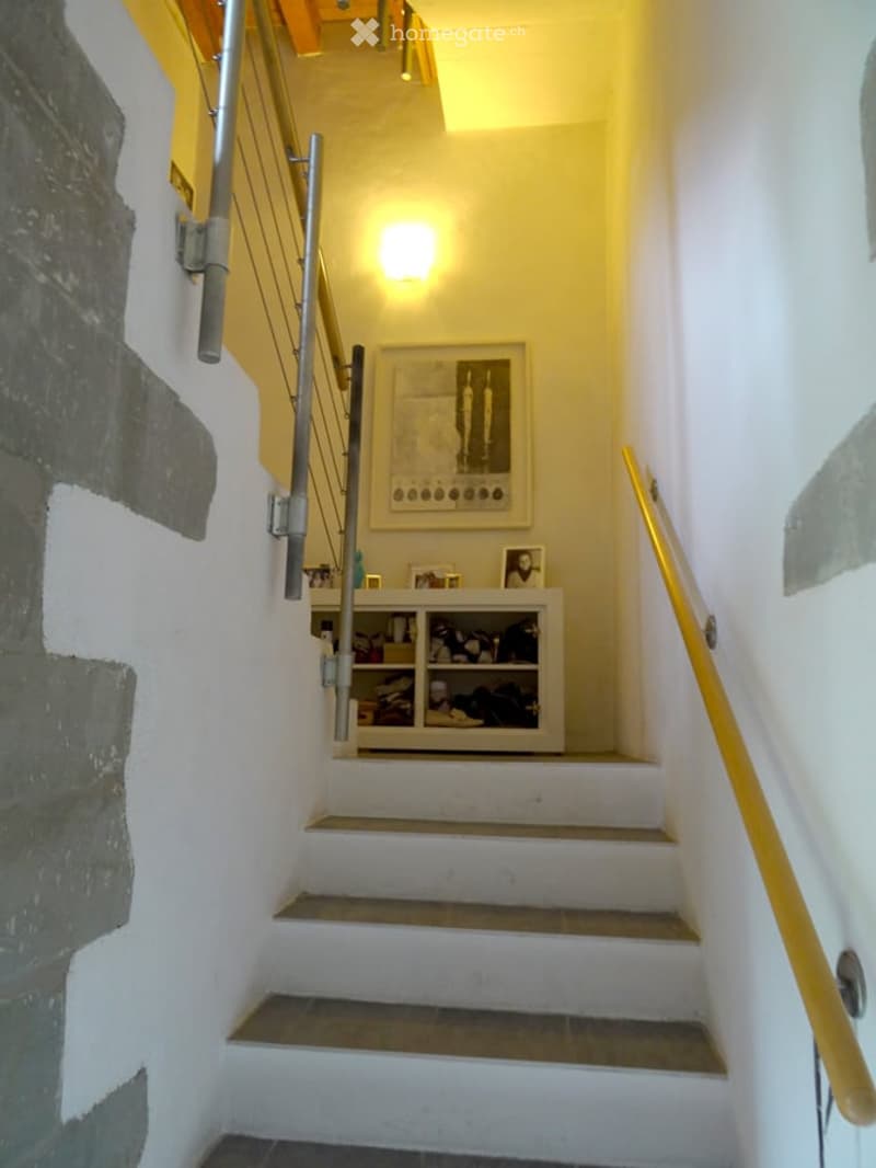 A narrow staircase with white walls, a metal railing on one side, and a wooden handrail on the other. A small shelf with various items is positioned against the wall, and a framed picture hangs above it. The lighting is warm, and the steps are tiled.