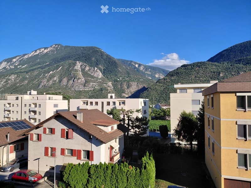 A multi-story residential building with red shutters and solar panels on the roof, surrounded by trees and parked cars, with a mountain range in the background.