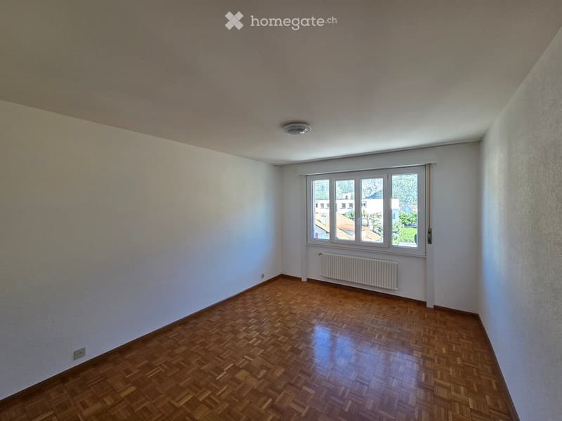 Empty room with wooden parquet flooring, white walls, and a large window with a radiator below it.