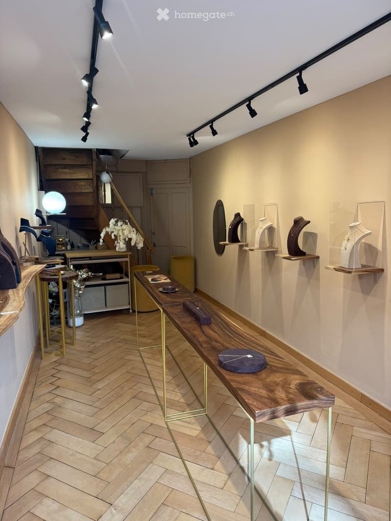 A well-lit hallway with wooden flooring, a long wooden table with decorative items, several small golden stools, shelves displaying necklaces, and a staircase leading upwards.