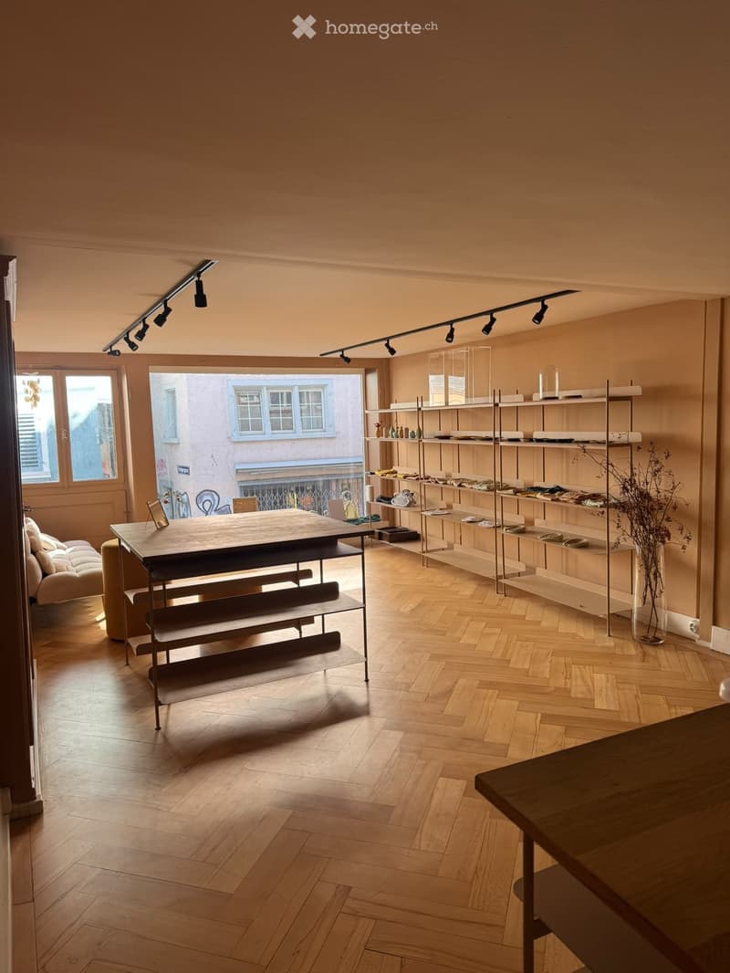 A modern, well-lit room with wooden flooring, a large window, a sofa, a table, shelves displaying items, and a vase with dried plants.