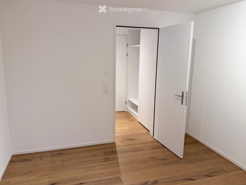 Empty room with light wooden flooring and white walls, featuring an open closet with a single shelf visible.