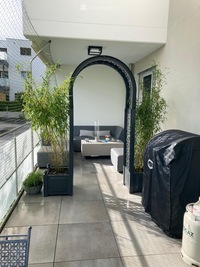 Modern outdoor patio with a sofa, coffee table, ottoman, and potted plants. Features a covered grill and a decorative archway.
