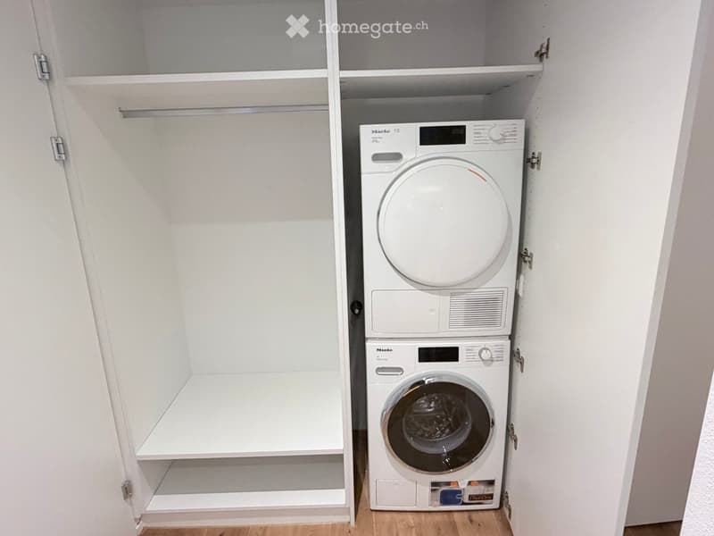 A small, well-organized laundry closet with white built-in shelving on one side and a stacked washing machine and dryer on the other.