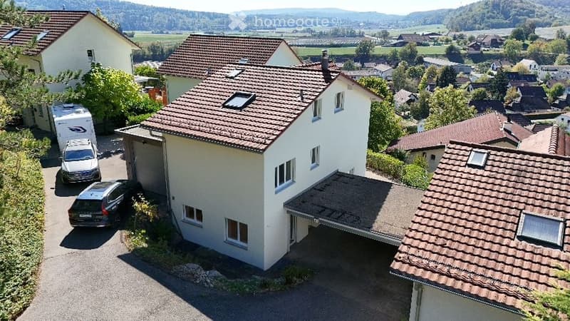 Aerial view of a two-story house with a red-tiled roof, white walls, and a garage. Several cars are parked in the driveway, and the surrounding area includes other houses and green fields.