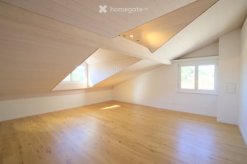 Empty attic room with wooden floors, white walls, and exposed wooden beams on the ceiling. Two windows provide natural light.