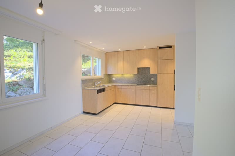 Modern kitchen with light wooden cabinets, tiled floor, and large windows overlooking a garden.