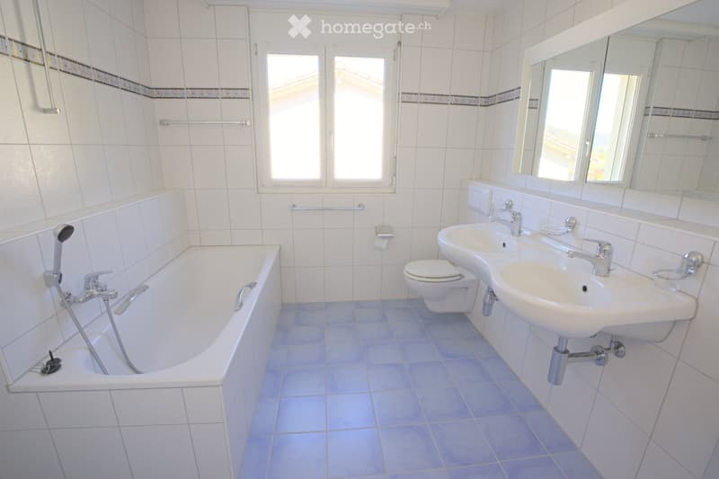 Modern bathroom with white tiles, a bathtub, a toilet, two sinks, and a large mirror above the sinks.