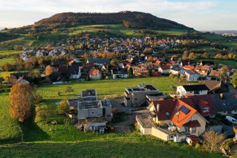 Aerial view of a suburban neighborhood with numerous houses, some featuring solar panels on their roofs, surrounded by green fields and trees.