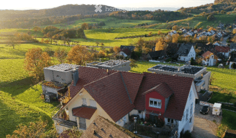 Aerial view of a residential building with a red-tiled roof, surrounded by green fields and scattered houses in the background.