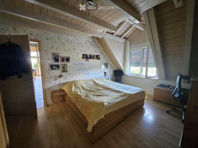 Bright, wooden-floored bedroom with slanted wooden ceiling beams, white brick accent wall, large windows with blinds, bed with yellow bedding, small wooden nightstand, framed pictures, and an office chair near the window.