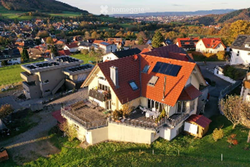 A modern two-story house with a red-tiled roof, large windows, and solar panels on the roof. The house features a spacious balcony and is surrounded by a well-maintained garden with trees and shrubs.