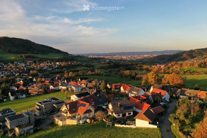 Aerial view of a residential area with numerous houses, red-tiled roofs, and surrounding green fields and hills.