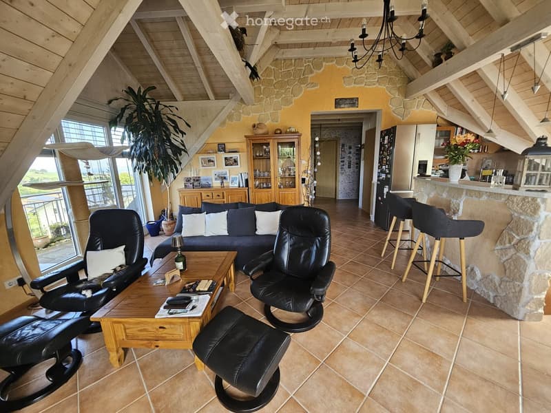 Bright living room with exposed wooden beams, a large window offering a view, a blue sofa with white cushions, a wooden coffee table, leather chairs, a bar area with stools, and a kitchen area visible in the background.