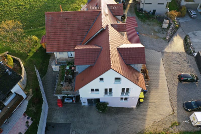 Aerial view of a two-story house with a red-tiled roof, white walls, and a garage. The house is surrounded by a paved driveway with several parked cars.