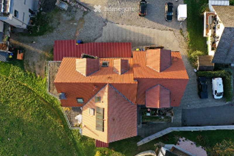 Aerial view of a modern two-story house with a red-tiled roof, multiple chimneys, and a garage. The house is surrounded by a paved driveway with several parked cars.
