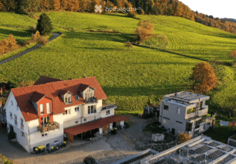 Aerial view of a large, modern house with a red-tiled roof, surrounded by green fields and trees. The house has multiple wings and a spacious driveway.