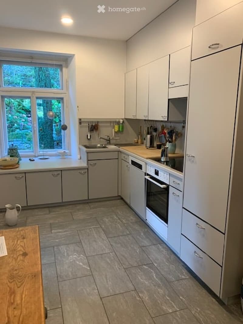 Modern kitchen with white cabinets, stainless steel appliances, and a large window overlooking greenery. Includes a sink, microwave, oven, countertop, and various kitchen utensils.