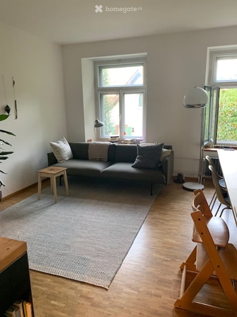 Modern living room with a grey sectional sofa, wooden coffee table, floor lamp, and a small plant. The room features light wooden flooring and large windows letting in natural light.