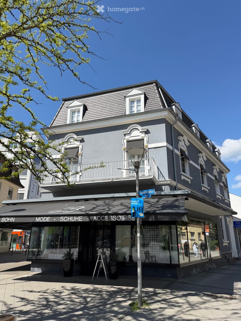 Three-story building with grey facade, white trim, and a balcony on the second floor. The ground floor features a storefront with large glass windows and a black awning displaying the text "Bohn Mode - Schuhe - Accessoires." The building has multiple wind