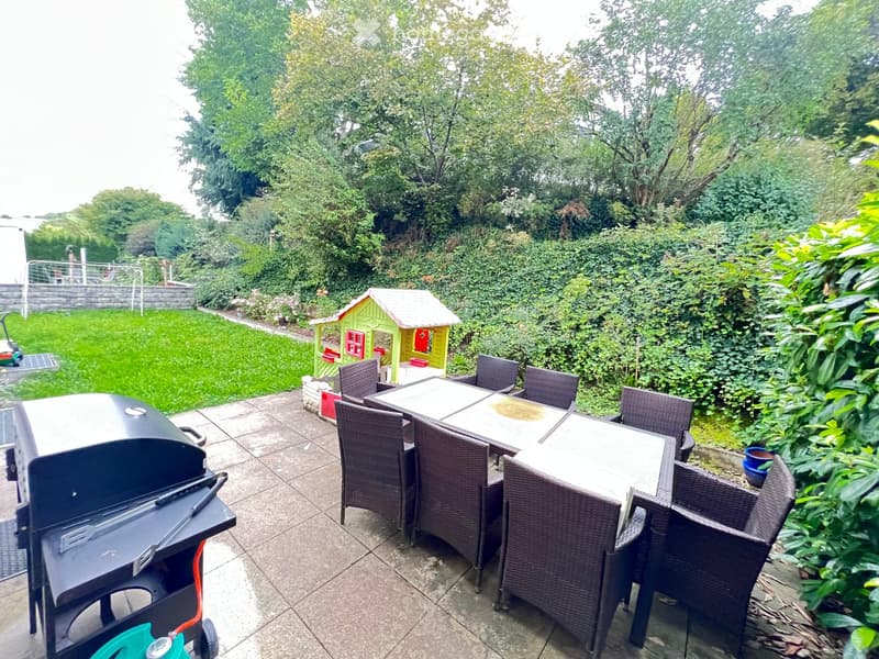 Outdoor patio with a round wicker table and chairs, a grill, and a small toy house in the background, surrounded by greenery.
