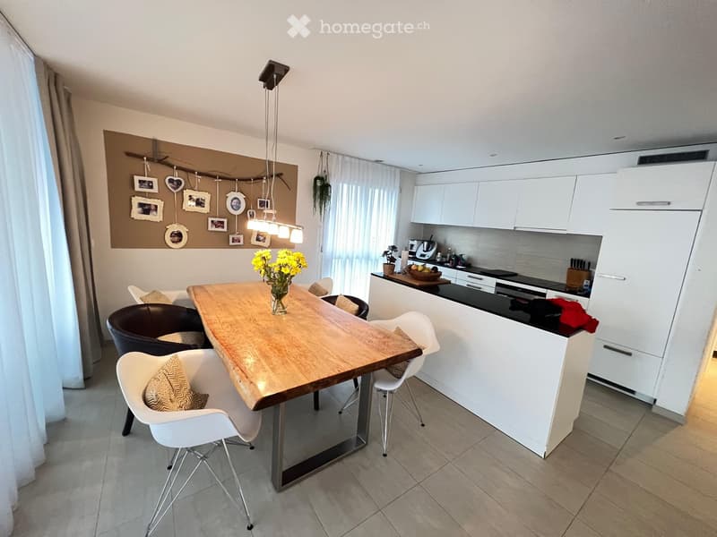 Modern kitchen with white cabinets, a wooden dining table, four chairs, and various appliances including a blender and oven. There is a flower vase on the table and picture frames on the wall.