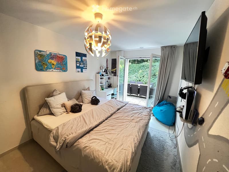 Cozy bedroom with a large bed, pillows, headboard, a chair, a small table, a screen, a decorative lamp, and a curtained window leading to a balcony.
