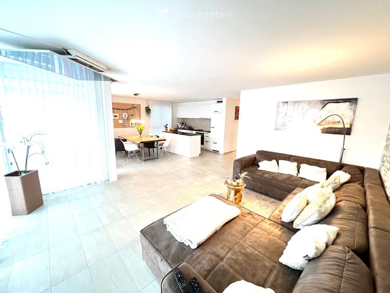Modern living room with a brown sectional sofa, coffee table, dining area with table and chairs, and a small kitchen area with white cabinets.