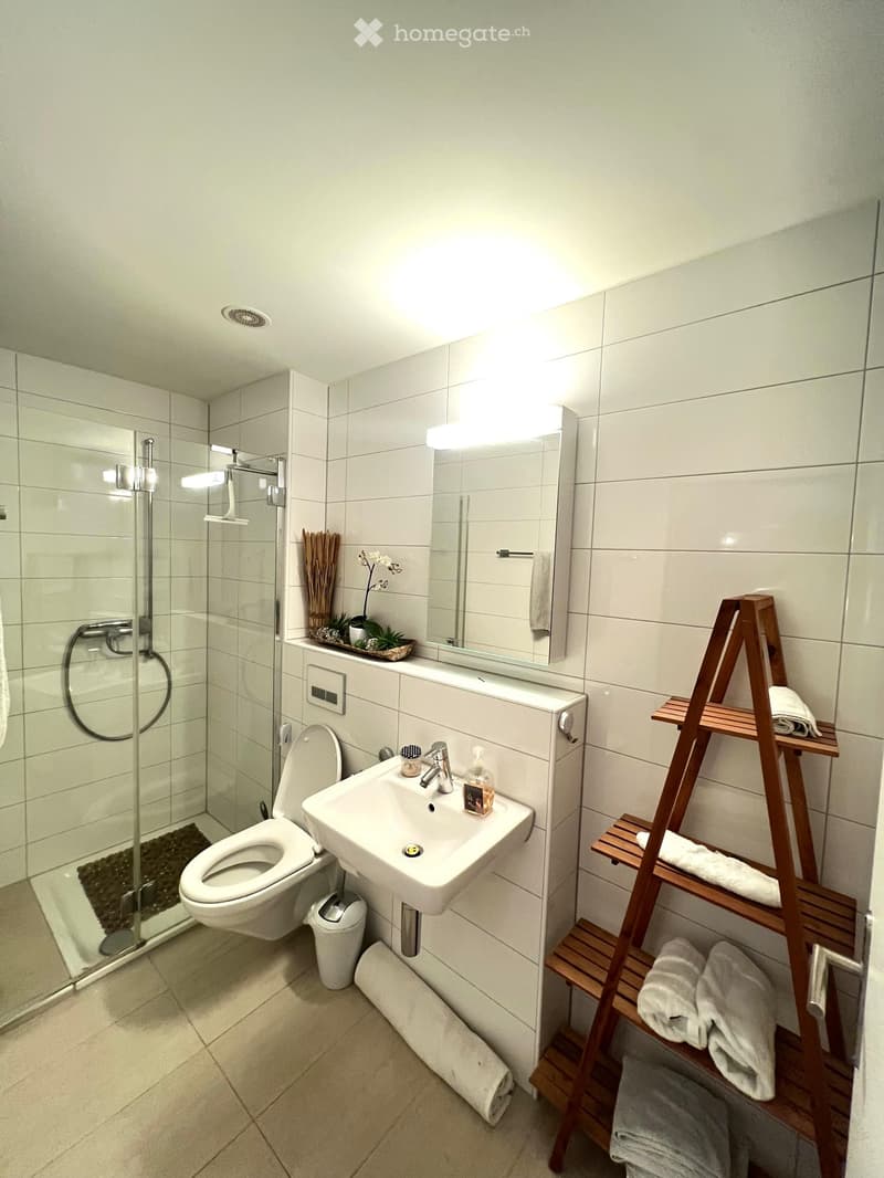 Modern bathroom with white tiles, a toilet, a sink with a mirror, a wooden towel rack, and a glass-enclosed shower area.