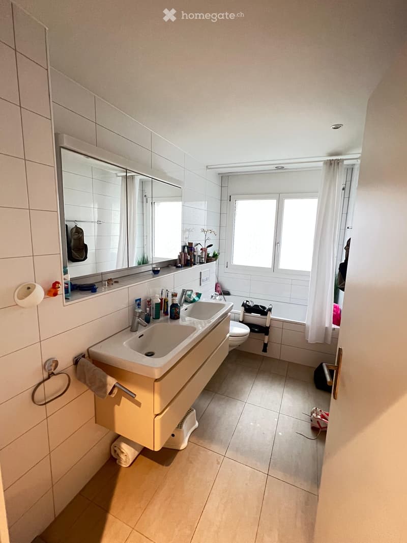 Modern bathroom with double sink, large mirror, toilet, bathtub, and window with curtain. Includes various toiletries and a small plant on the sink counter.