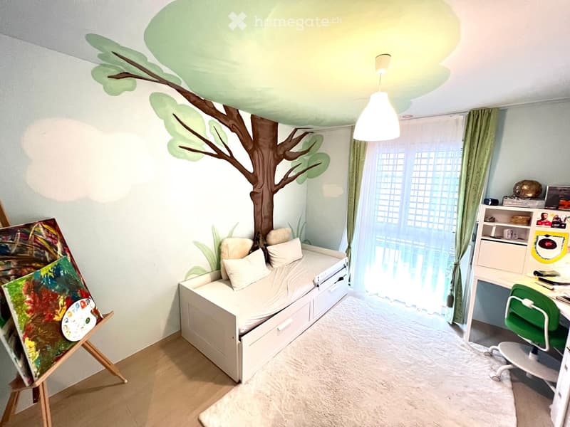 Cozy bedroom with a white bed, desk, green chair, and an easel with paintings. Features a mural of a tree on the wall and large windows with curtains.