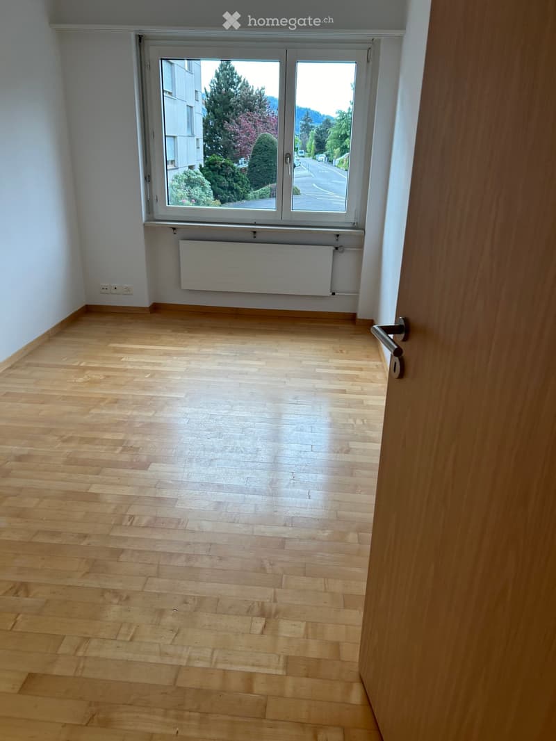 Empty room with wooden flooring, white walls, large window with view of greenery, radiator, and door handle visible.