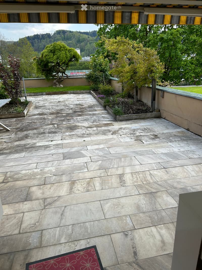 Outdoor patio with stone tiles, small garden beds, trees, and a scenic view of a hilly landscape.