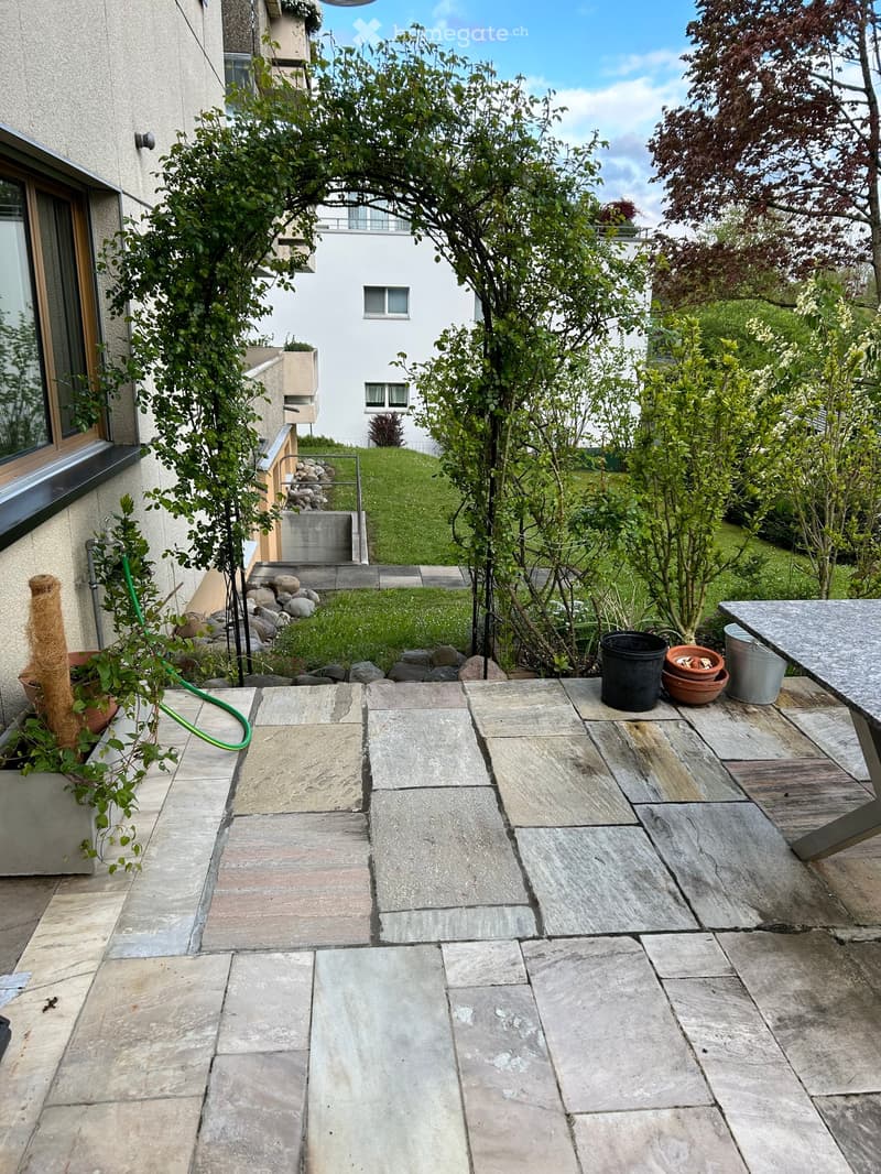Garden area with stone-paved patio, small grassy lawn, trees, and potted plants.