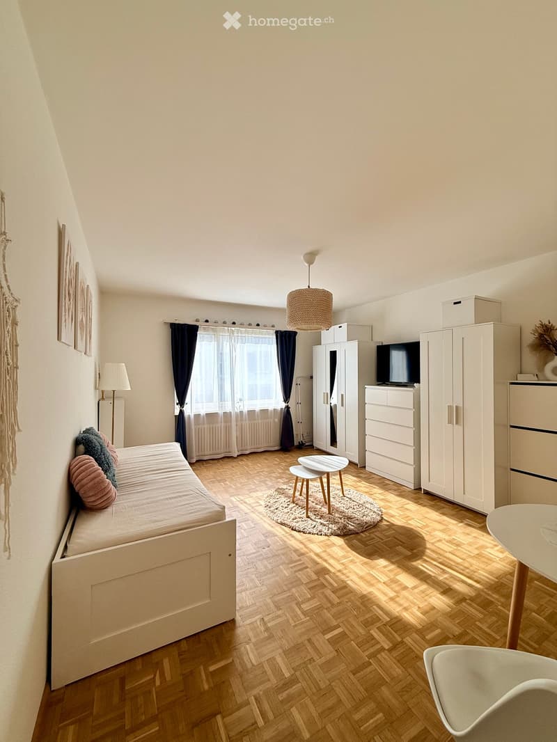 Cozy bedroom with a wooden bed, small table, chair, wardrobe, and a hanging lamp. The room features wooden flooring, a window with curtains, and decorative elements like a picture frame and a woven wall hanging.