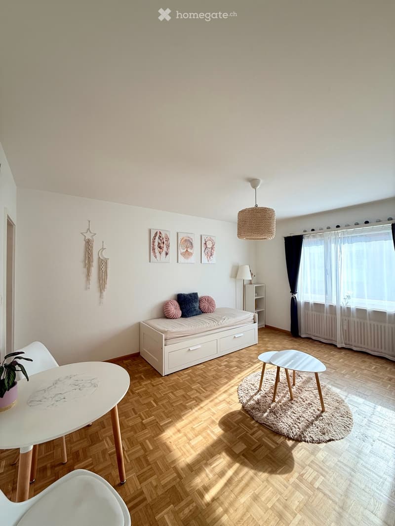 A modern, minimalist bedroom with a bed, a small table, two chairs, a round stool, a hanging lamp, and decorative items including a plant and picture frames.